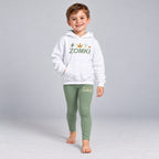 Visualización editorial generada con IA con el mismo modelo infantil sintético del conjunto ZOMKI Corona Verde salvia, en una pose nueva de paso frontal. Sudadera blanca y leggings coordinados con rayo y trazos de energía. No representa a una persona real.
