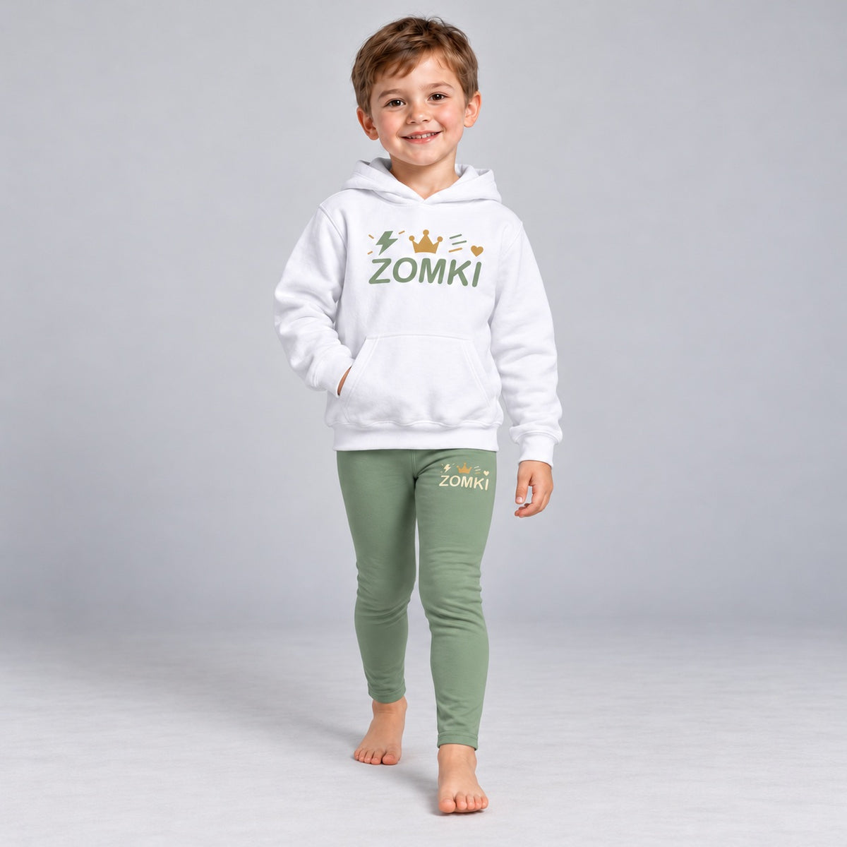 Visualización editorial generada con IA con el mismo modelo infantil sintético del conjunto ZOMKI Corona Verde salvia, en una pose nueva de paso frontal. Sudadera blanca y leggings coordinados con rayo y trazos de energía. No representa a una persona real.