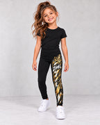 Leggings infantiles negros ZOMKI Graffiti Dorado con modelo sintético IA niña; estampado blanco y dorado con corona y corazón sobre la i en la pierna izquierda