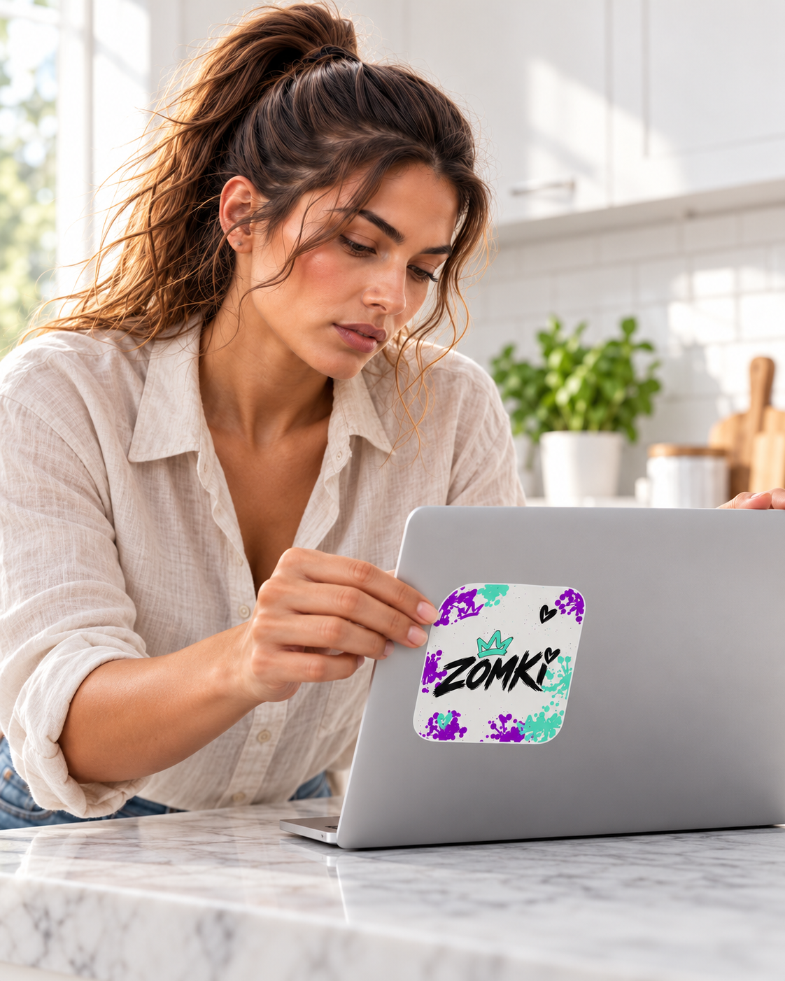 Modelo oficial ZOMKI aplicando la pegatina individual Mint Violet Blanco a un portátil de atrezo en una cocina luminosa.